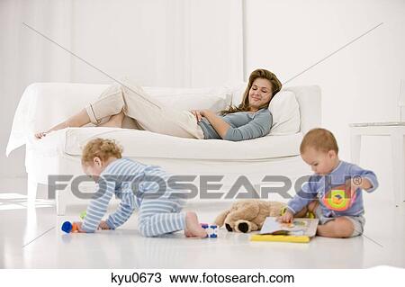 Stock Image - Babies playing with toys and their mother resting on a couch. Fotosearch
