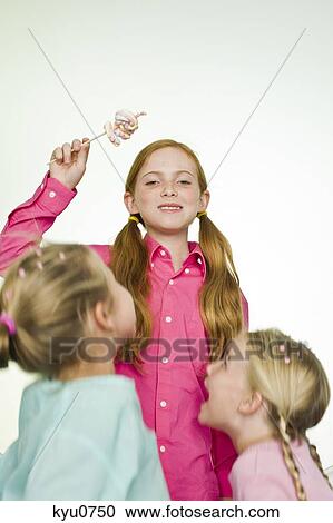Stock Image - Girl holding a lollipop with her two friends standing beside her. Fotosearch
