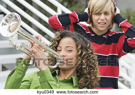 Girl playing a trumpet with her friend covering his ears in the background View Large Photo Image Stock Photo - Girl playing a trumpet with her friend covering his ears in the background. Fotosearch