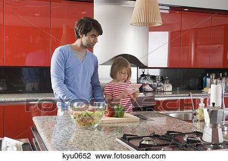 Stock Image - Girl playing a video game with her father chopping bell pepper beside her. Fotosearch