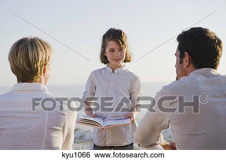 Girl reading a book in front of her parents View Large Photo Image Stock Photograph - Girl reading a book in front of her parents. Fotosearch