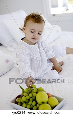 Picture - Little girl sitting on a bed, taking grapes. Fotosearch