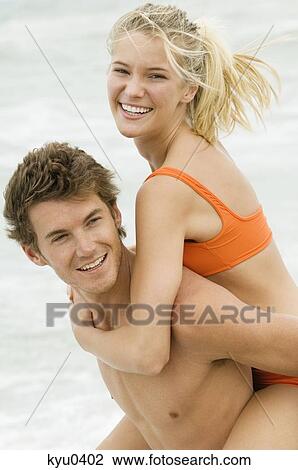 Stock Image - Man giving woman piggyback ride on the beach. Fotosearch