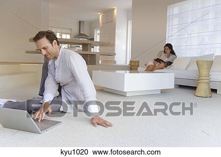 Stock Image - Man working on a laptop with a woman reading a book in the background. Fotosearch
