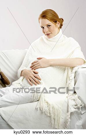 Stock Image - Portrait of a pregnant young woman sitting on a couch and smiling. Fotosearch