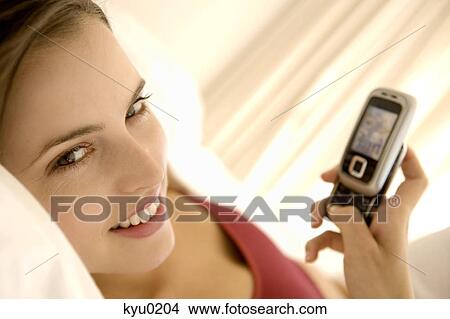 Picture - Portrait of a young woman lying on a bed, holding her cell. Fotosearch
