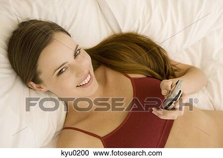 Portrait of a young woman lying on a bed, holding her cell View Large Photo Image Stock Image - Portrait of a young woman lying on a bed, holding her cell. Fotosearch