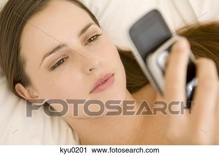 Stock Image - Portrait of a young woman lying on a bed, holding her cell. Fotosearch