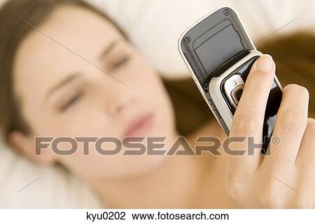 Stock Image - Portrait of a young woman lying on a bed, holding her cell. Fotosearch