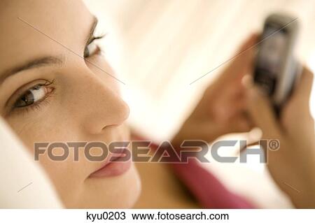 Stock Image - Portrait of a young woman lying on a bed, holding her cell. Fotosearch