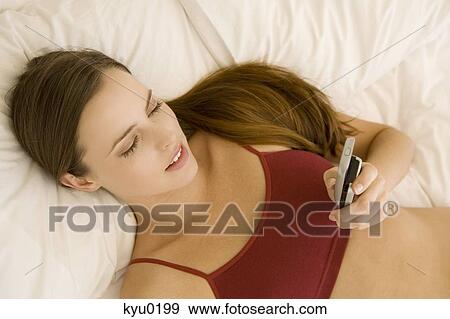 Stock Photo - Portrait of a young woman lying on a bed, holding her cell. Fotosearch