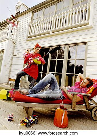 Stock Photograph - Youg woman holding baby and feather duster, shouting at man lying on a deck chair. Fotosearch