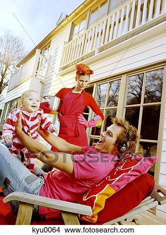 Picture - Young man playing with baby lying on a deck chair, angry woman in background. Fotosearch