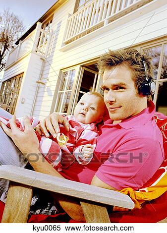 Stock Photography - Young man with headphones holding baby, lying on a deck chair. Fotosearch