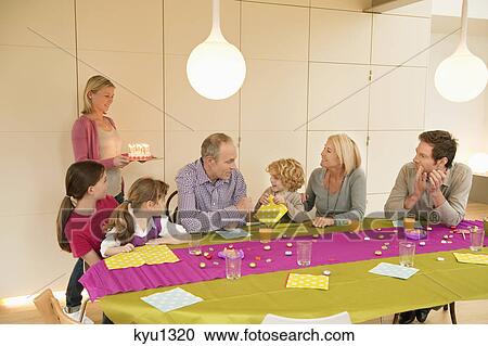 Stock Image - Family at a birthday celebration with a woman bringing cake in the background. Fotosearch