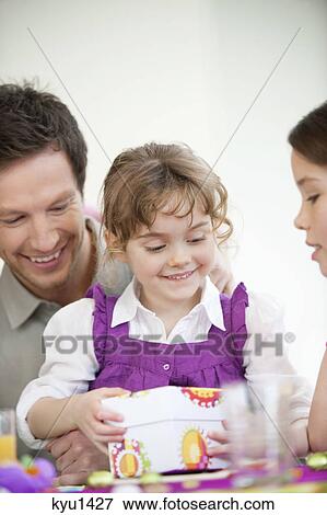 Stock Photo - Girl opening her birthday present with her father and sister. Fotosearch