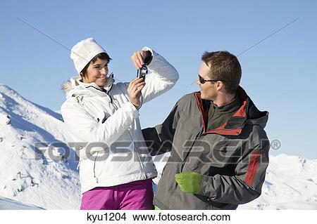 Picture - Young woman in ski wear taking a photo of her boyfriend. Fotosearch