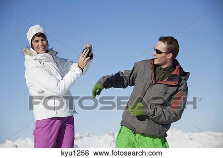 Stock Photo - Young woman in ski wear taking a photo of her boyfriend. Fotosearch