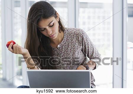 Stock Image - Beautiful woman holding an apple while using a laptop. Fotosearch