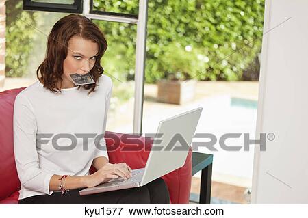 Stock Photo - Young woman holding credit card in mouth, using laptop. Fotosearch