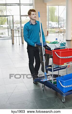 Stock Image - Male cleaner pushing trolley with cleaning equipment. Fotosearch