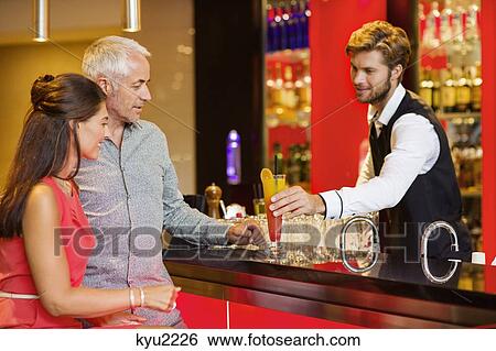 Stock Photograph - Bartender serving drink to a couple at bar counter. Fotosearch