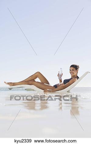 Stock Photo - Beautiful woman holding a water bottle while sunbathing on the beach. Fotosearch