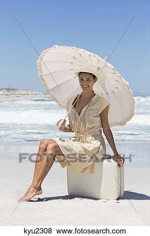 Stock Photo - Beautiful woman sitting on a suitcase with an umbrella on the beach. Fotosearch