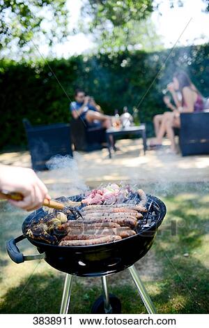 Close up of a barbecue grill with meat and sausages cooking during a summer garden party with people in background. View Large Photo Image Stock Image - Close up of a barbecue grill with meat and sausages cooking during a summer garden party with people in background.. Fotosearch