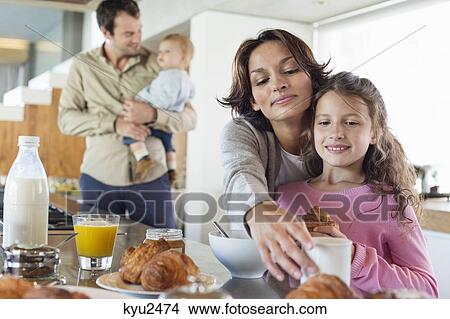Picture - Girl having breakfast beside her mother at a kitchen counter. Fotosearch