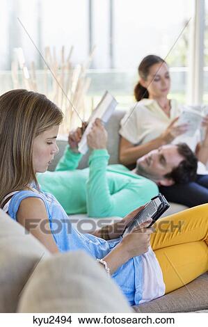 Picture - Girl using a digital tablet with her parents reading books at home. Fotosearch