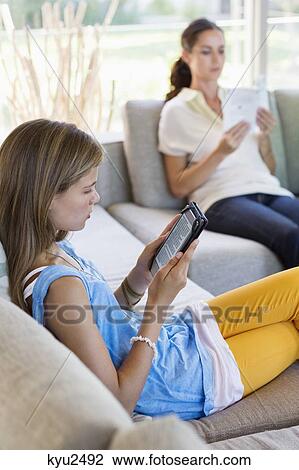 Stock Image - Girl using a digital tablet with her mother reading book at home. Fotosearch