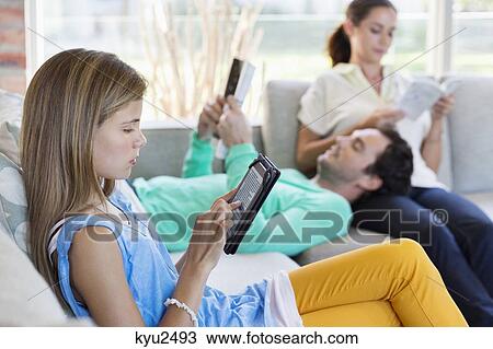 Stock Image - Girl using a digital tablet with her parents reading books at home. Fotosearch