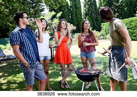 Stock Photograph - Groupe of happy and cheerful young people having a toast around barbecue grill during summer holiday party outdoor in the garden.. Fotosearch