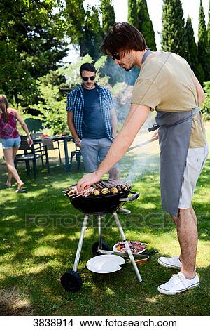 Picture - Handsome young man cooking meat in a barbecue party outdoor in the garden during summer holiday.. Fotosearch