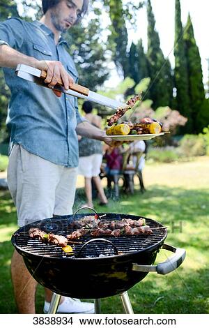 Picture - Handsome young man cooking meat in a barbecue party outdoor in the garden during summer holiday.. Fotosearch