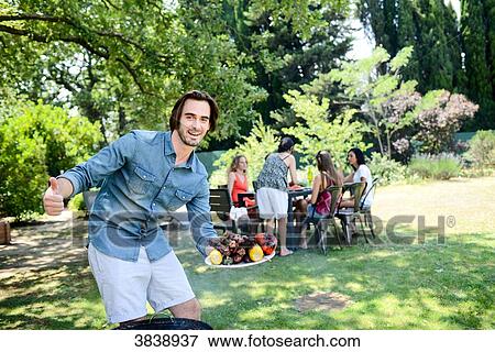 Stock Photo - Handsome young man showing grilled meat in barbecue party outdoor in the garden during summer holiday.. Fotosearch