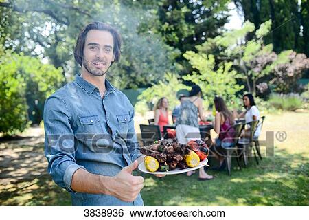 Handsome young man showing grilled meat in barbecue party outdoor in the garden during summer holiday. View Large Photo Image Stock Photograph - Handsome young man showing grilled meat in barbecue party outdoor in the garden during summer holiday.. Fotosearch