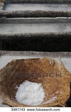 Stock Image - Indonesia, Bali, salt harvesting. The salt producer takes sea water with 2 baskets. She shakes the baskets so that the water is poured on the sand. After evaporation, the sand is then removed, and put in a half coconut tree trunks. Fotosearch