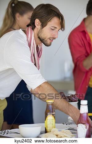 Stock Image - Man arranging a dining table with his friends in the background. Fotosearch