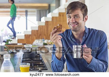 Stock Photo - Man having breakfast at a kitchen counter with his wife in the background. Fotosearch