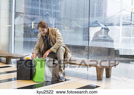 Stock Image - Man sitting at an airport lounge looking at shopping bags. Fotosearch