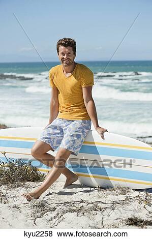 Stock Photo - Man sitting on a surfboard on the beach. Fotosearch