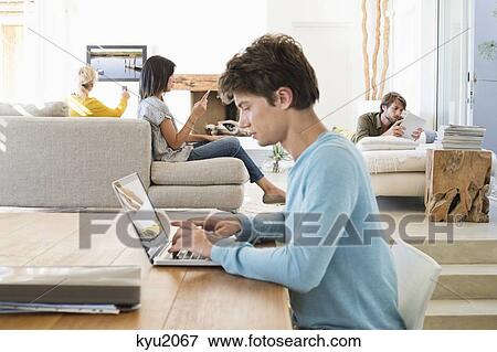 Stock Photo - Man using a laptop with his friends using electronic gadgets in background. Fotosearch
