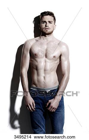 Picture - Topless man against a white wall; wearing jeans, serious, hard shadow on the wall, looking at the camera, top botton of his jeans undone. Fotosearch