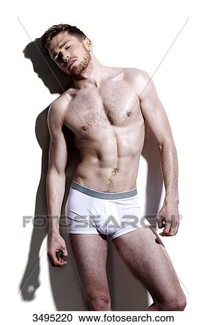 Stock Image - Topless man, against a white wall; wearing white pants, serious, hard shadow on the wall, head back, eyes closed, arms alongside his body. Fotosearch