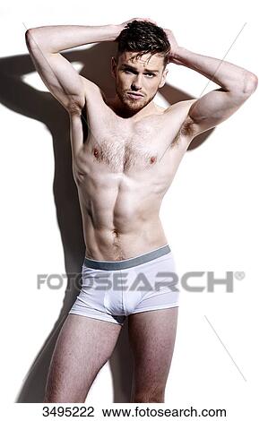 Stock Image - Topless man, against a white wall; wearing white pants, serious, poised, hard shadow on the wall, looking at camera, frowning, hands behind the head, arms up.. Fotosearch