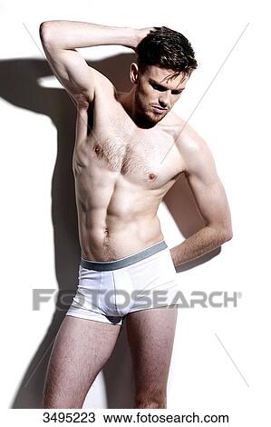 Stock Image - Topless man, against a white wall; wearing white pants, serious, hard shadow on the wall, looking down, arm up, one hand behind the head. Fotosearch