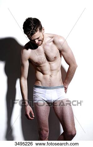 Stock Photograph - Topless man, against a white wall; wearing white pants, hard shadow on the wall, looking down smiling.. Fotosearch