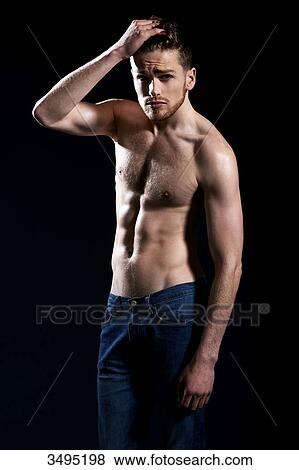 Stock Photo - Topless man, black background; one hand in his hair, arm up, wearing jeans, serious, forehead and eyebrows creased.. Fotosearch